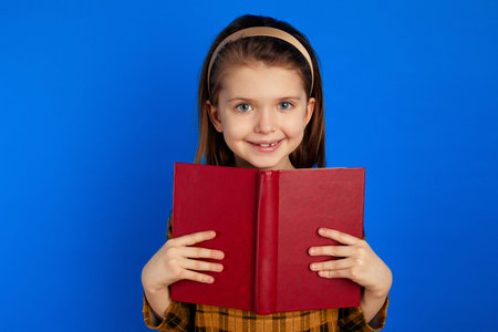 Little kid has charming smile, wears checkered dress, carries red book