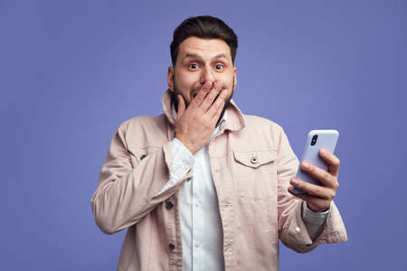 Shocked man covers mouth with hand, reads horrified news from smartphoneの写真素材