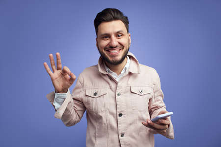 Handsome man smiling and gesturing ok sign while holding mobile phoneの写真素材