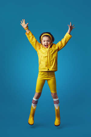 Full body excited kid girl in yellow raincoat and boots smiling and jumpingの写真素材