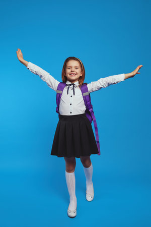 Schoolgirl wearing uniform and backpack, raises hands up against blue wall