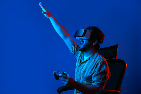 Young bearded man in VR glasses screams and celebrates winning in virtual game, sitting on chair and...