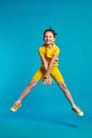 Funny child girl wears yellow sportswear and pedometer band, jumping on a colored blue studio...