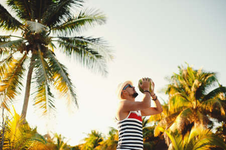 Young attractive man wearing sunglasses and straw hat drinking coconut milk on the tropical beach on sunny summer day during holidays vacationの写真素材