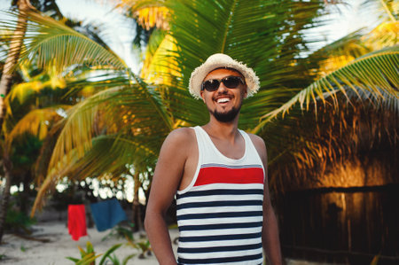 Young bearded tourist man wearing sunglasses and straw hat smiling broadly against background of palm treesの写真素材