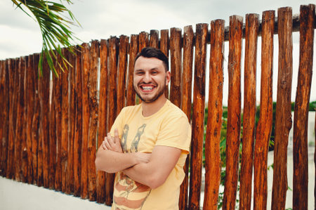 Confident smiling bearded young man in yellow t shirt with folded arms, smiling and leaning against...