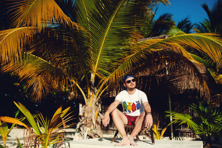 Attractive young tourist man in white t shirt and sunglasses rests sitting on chair and looking away...