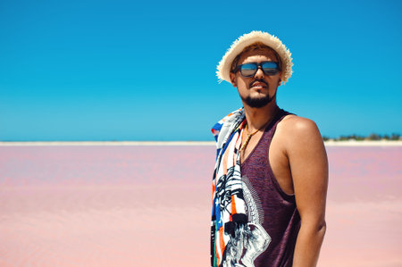 Close up portrait of young serious man in sunglasses and straw hat posing against pink salt lake in...