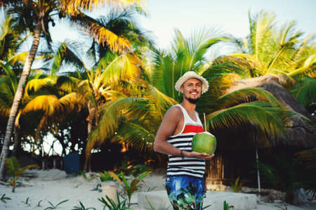 Handsome happy tourist man in tshirt and straw hat holding coconut on the tropical beach on sunny summer day during holidays vacationの写真素材