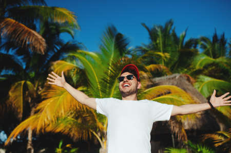 Cheerful attractive boy in white t shirt and sunglasses, gesturing triumph with raised hands against palm trees backgroundの写真素材