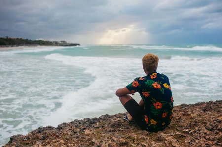Back view of adult man sitting on the edge of the abyss facing to the sea with big waves, alone depressed person, anxietyの写真素材
