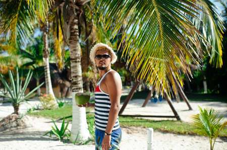 Attractive tourist man wearing sunglasses and straw hat holding coconut on the tropical beach on sunny summer day against palm treesの写真素材