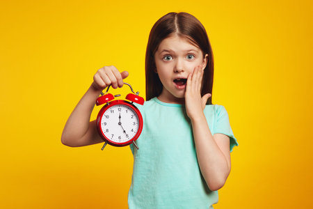 Young little girl kid holding classic bell alarm clock over isolated yellow background, shocked for mistake, expression of fear, scared in silenceの写真素材