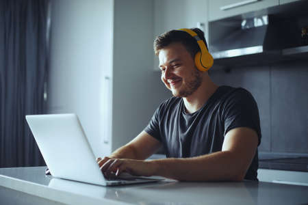 Smiling young man with yellow headphones using laptop studying online, working from home. Happy guy typing on pc notebook, surfing internetの写真素材
