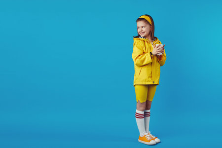 Full body photo of little kid wears raincoat and yellow headband, holds takeaway cup with hot...