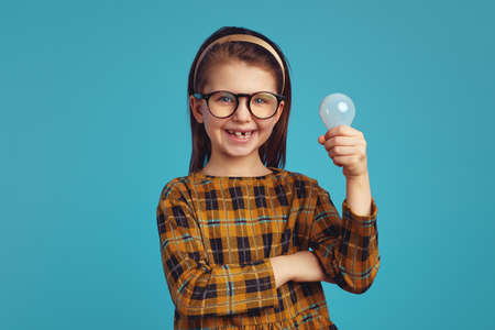 Attractive cheerful little kid with light bulb smiling while having brilliant idea against vivid blue backgroundの写真素材