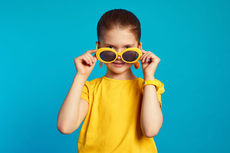 Lovely little girl in yellow shirt, looking at camera while adjusting sunglasses, isolated over blue...