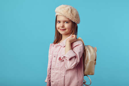 Cute beautiful little girl in pink jacket smiling while holding backpack in hand against blue background, free empty space for textの写真素材