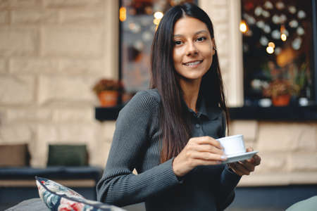 Cheerful young woman with a cup of coffee sitting at cafe. Female having coffee at coffee shopの写真素材