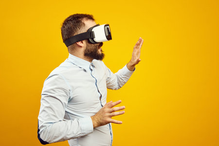 Side view of young bearded man wearing virtual reality helmet touching the air, isolated over yellow...