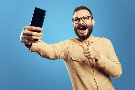 Happy caucasian male wearing eyeglasses and beige shirt, smiling and showing thumb up gesture while taking selfie against blue backgroundの写真素材