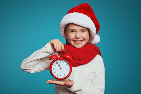 Adorable kid wearing santa hat and red scarf, holding red alarm clock on a blue backgroundの写真素材