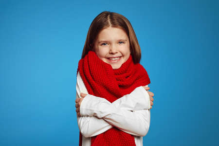 Little girl kid feels freezing, crosses arms over body, trembles from cold, wears red scarf, had walk during frosty weather, isolated over blueの写真素材