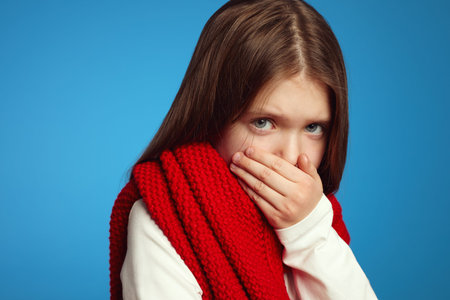 Close up photo of cute girl kid in red scarf sneezing or coughing covering mouth with palm isolated...