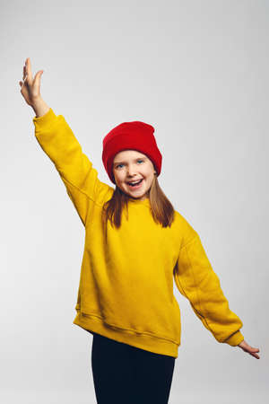 Vertical photo of laughing girl in yellow hoodie and red hat holding hands up in gesture of victory on white backgroundの写真素材