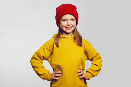 Confident hipster little kid in yellow hoodie and red hat holds hands on waist and smiles broadly, poses with happy expression over white backgroundの写真素材