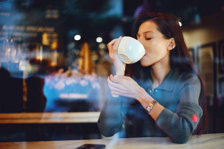 Young adorable female drinking coffee while sitting inside a coffee shopの写真素材
