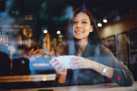 Smiling woman holding a coffee cup close to her face sitting inside a coffee shop. Young cute girl drinking coffeeの写真素材