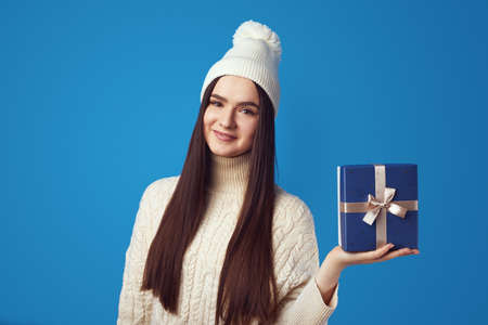 Happy young female wearing white knitted oversize sweater and hat, holding wrapped present box for christmas, isolated over blue backgroundの写真素材