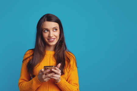 Dreamy young woman holds mobile phone has thoughtful expression thinks about received message looks away isolated over blue backgroundの写真素材