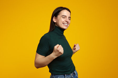 Side view of young girl in green t-shirt doing winner gesture with closed eyes, posing isolated on yellow backgroundの写真素材