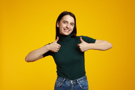 Portrait of happy young woman isolated over yellow background showing thumbs up gesture while smiling broadly, wearing green t-shirtの写真素材