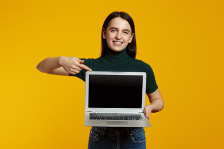 Trendy hipster girl wearing green t-shirt pointing at new laptop in hands, isolated over yellow background.の写真素材