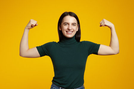 Strong powerful woman with toothy smile, raises arms and shows biceps, wears casual green t shirt, isolated over yellow backgroundの写真素材
