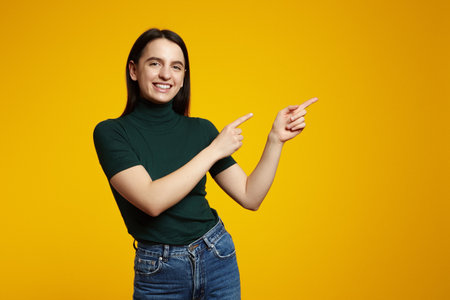 Portrait of young attractive happy positive good mood girl smiling and pointing finger to copy space isolated on yellow color backgroundの写真素材