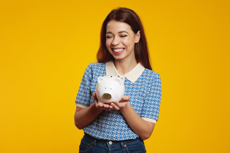 Young woman in casual blue shirt holding white piggy bank with lots of money while smiling broadly with closed eyes against yellow backgroundの写真素材