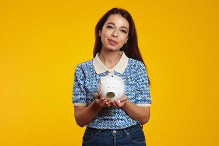 Cute girl in casual blue shirt holding white piggy bank with lots of money isolated over yellow backgroundの写真素材