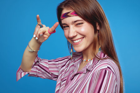 Close up portrait of excited young hippie woman smiling in striped pink shirt showing rock and roll...