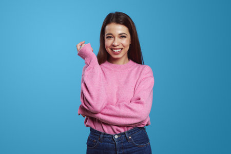 Optimistic young ethnic female model with brunette hair smiling happily and looking at camera against blue backgroundの写真素材