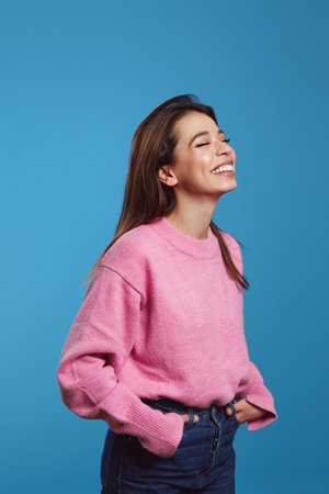 Vertical photo of cheerful young woman model standing in pink jumper and blue jeans, smiling happily with closed eyes, isolated over bright blue backgroundの写真素材