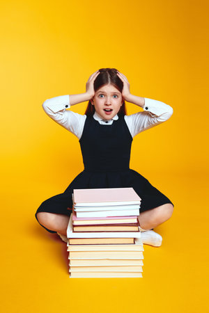 Vertical photo of astonished girl in school uniform keeping hands near head while sitting near stack of books, isolated over yellow backgroundの写真素材