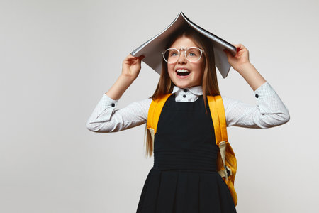 Surprised schoolgirl with eyeglasses wearing school uniform and yellow backpack yelling and covering head with book against white backgroundの写真素材
