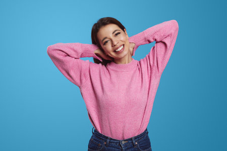Young ethnic female model in blue jeans and pink pullover holding hands behind head and happily smiling against blue backgroundの写真素材