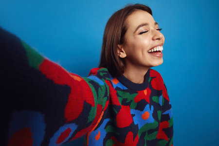 Overjoyed attractive hispanic girl smiling with closed eyes while making selfie, wearing colorful sweater, isolated over blue backgroundの写真素材