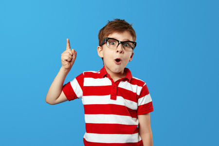 Smart amazed little boy wearing striped red polo shirt and eyeglasses opening mouth and pointing up while having idea against blue background. Child studio portraitの写真素材