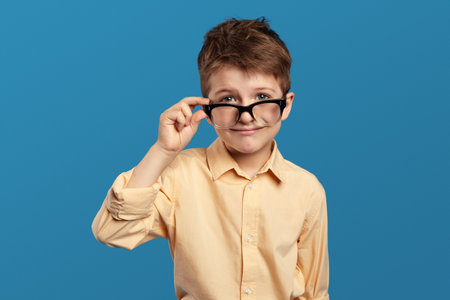 Close up portrait of sad, worried little kid boy in beige shirt, looking stressed at camera through glasses, isolated over blue backdrop. Negative human emotions, facial expressions, feelingsの写真素材